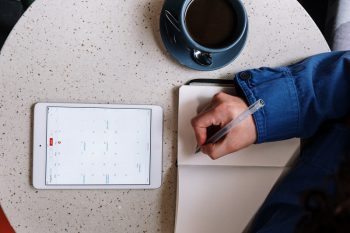 person's arm writing in a journal with a calendar from a tablet