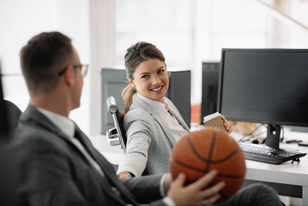 Sports management professionals interact at their desks Two sports management workers interact at their desks as the smiling woman reaches over to the man, who is holding a basketball.