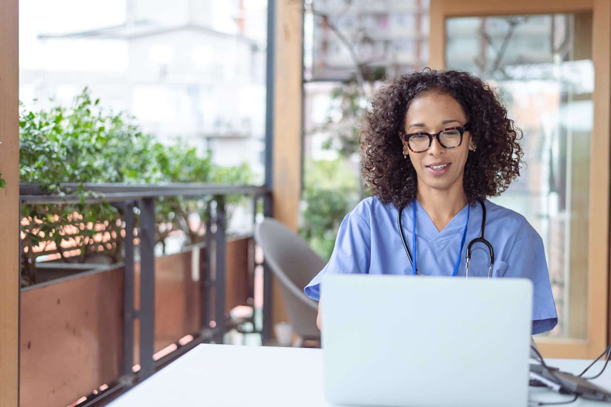 Nurse in blue scrubs with a stethoscope working on a laptop at an outdoor table near a leafy patio.