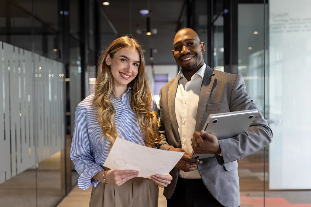 Two business professionals holding a report and a laptop in an office hallway